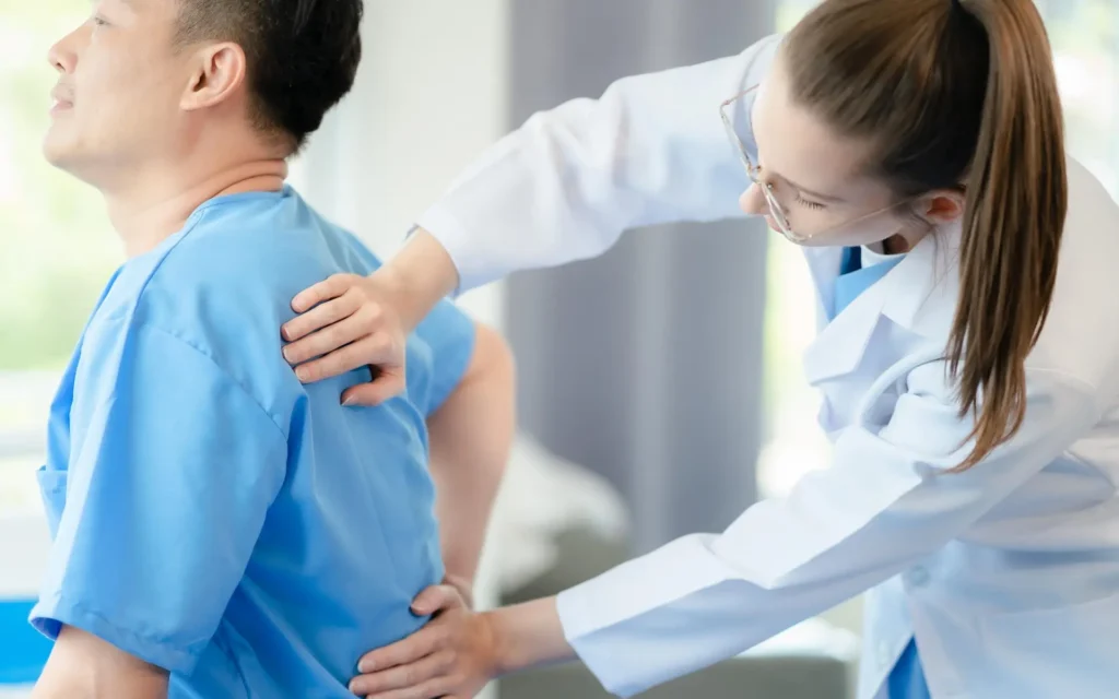A healthcare professional in a white coat is examining the back of a patient wearing blue scrubs. The patient is sitting and holding their lower back, while the healthcare professional uses both hands to palpate the patient's upper back.