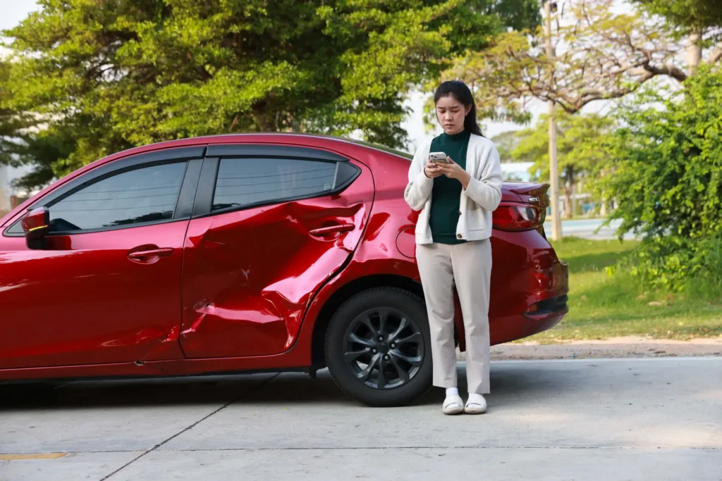Person standing next to a red car with a dented side, looking at their phone