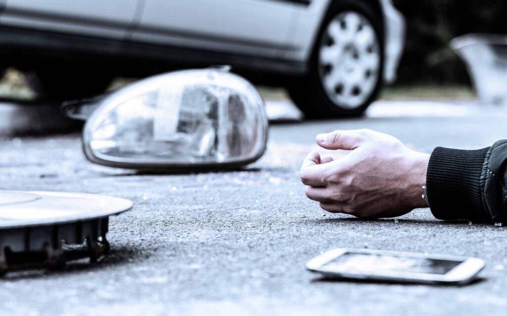 Close-up of a person's hand lying on the ground beside a smartphone, with a car headlight and part of a vehicle in the background, suggesting an accident or emergency
