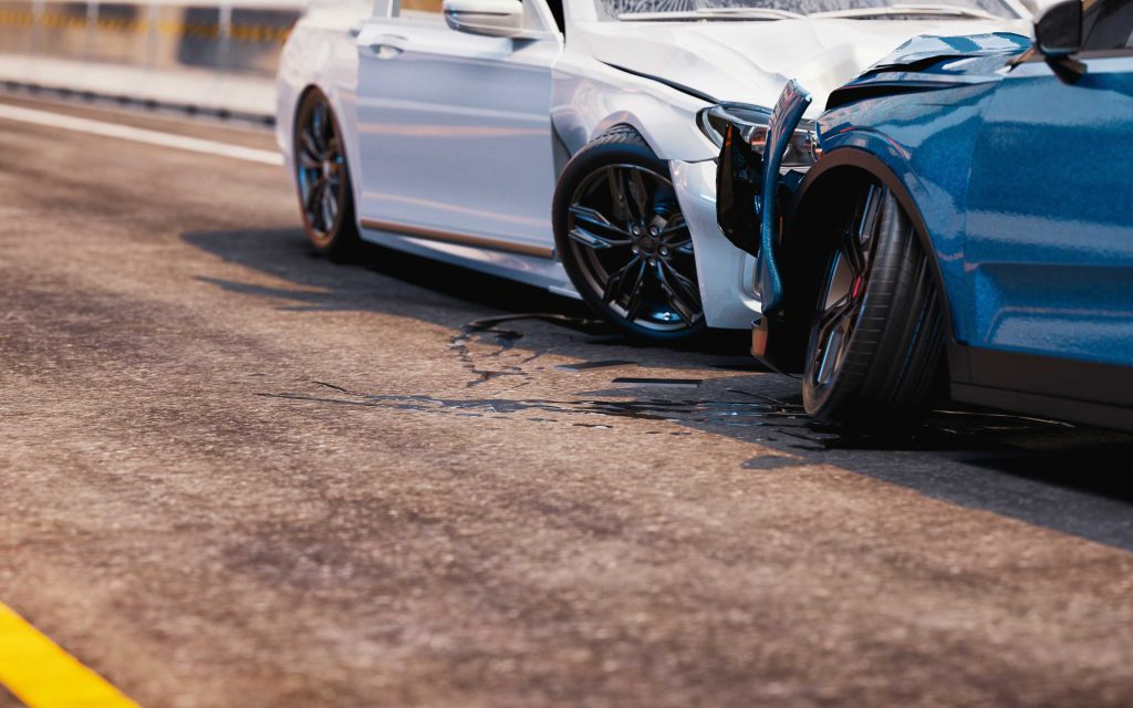 Two-car accident on a paved road, with a white car rear-ending a blue car, showing visible damage and fluid spill beneath both vehicles.