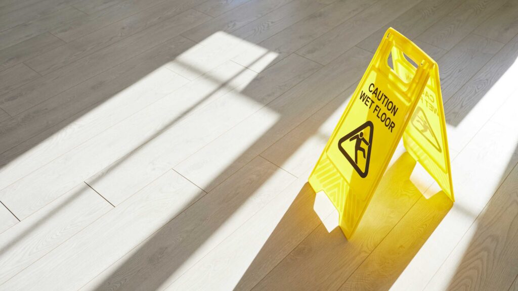 Yellow caution wet floor sign placed on light wooden flooring in sunlight.