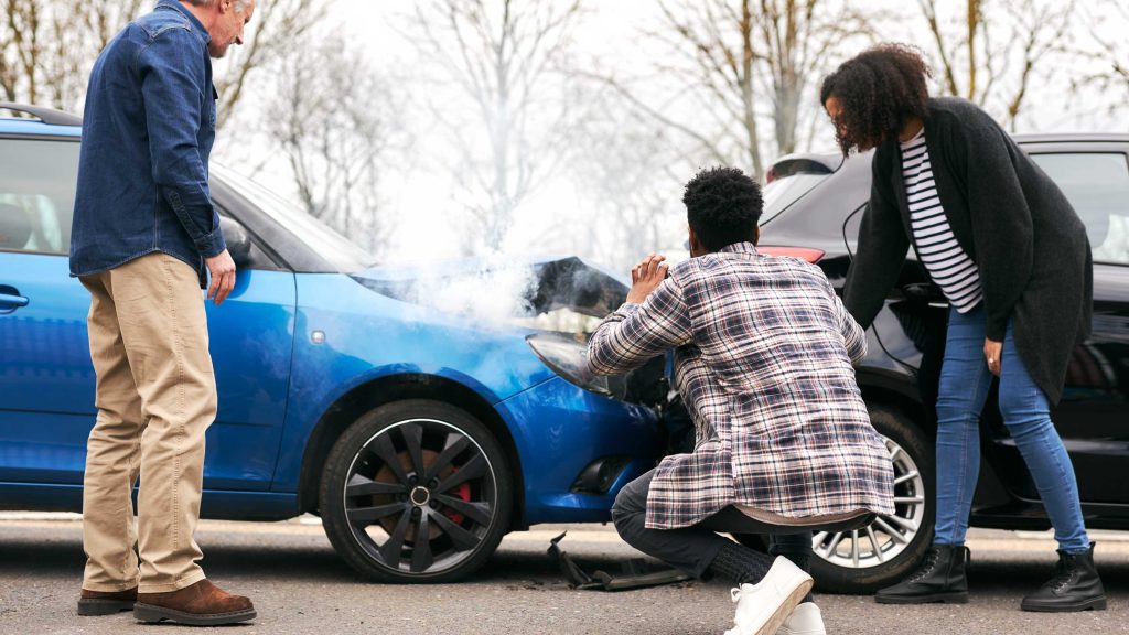 Three people inspecting two cars after a front-end collision with smoke rising from the engine.