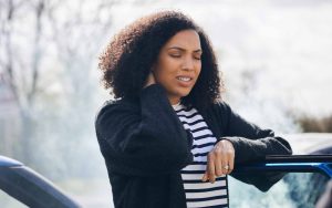 Woman standing beside a car holding her neck in pain after an accident, showing signs of discomfort or injury.