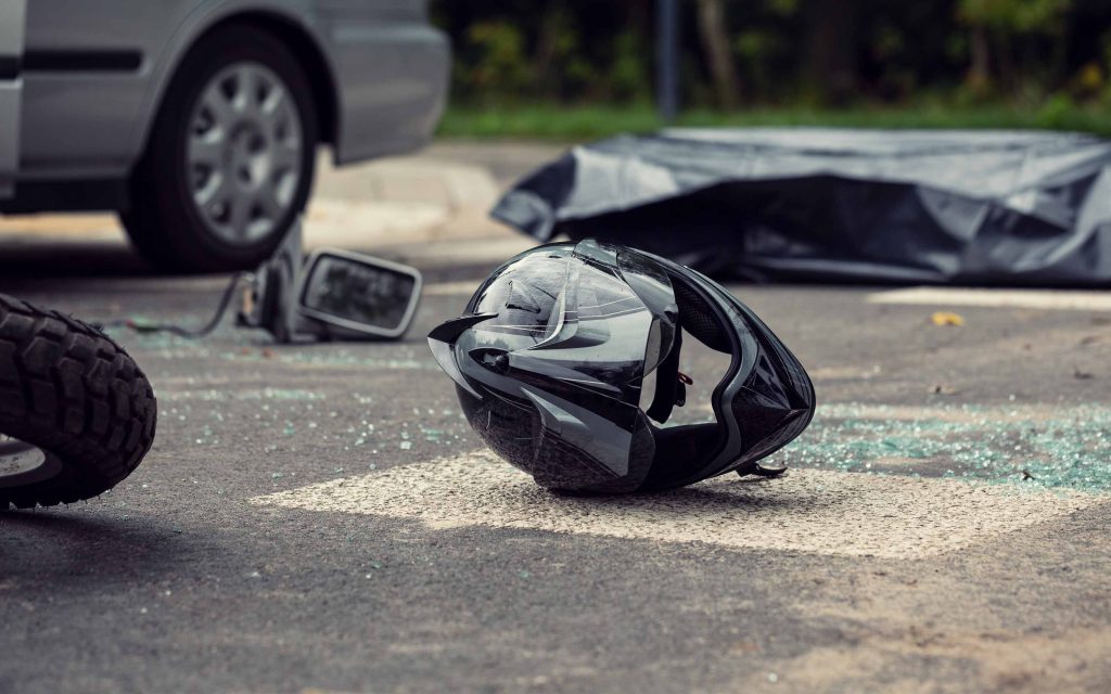 Motorcycle accident scene showing a damaged helmet on the road near a car and broken glass.