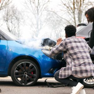 Three people inspecting two cars after a front-end collision with smoke rising from the engine.