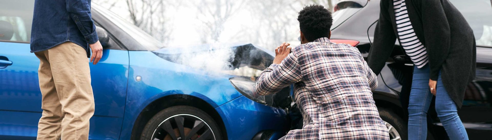 Three people inspecting two cars after a front-end collision with smoke rising from the engine.