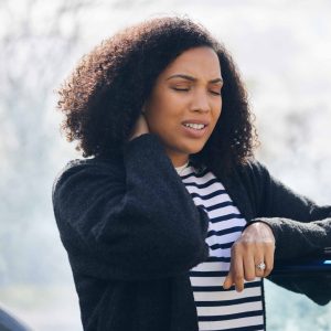 Woman standing beside a car holding her neck in pain after an accident, showing signs of discomfort or injury.