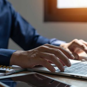 A man is typing on a laptop in front of a window. The laptop is open and the man's hands are on the keyboard. There are several notebooks and a tablet on the desk