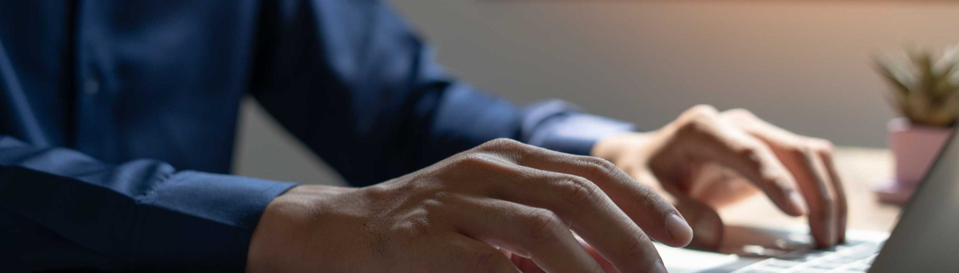 A man is typing on a laptop in front of a window. The laptop is open and the man's hands are on the keyboard. There are several notebooks and a tablet on the desk