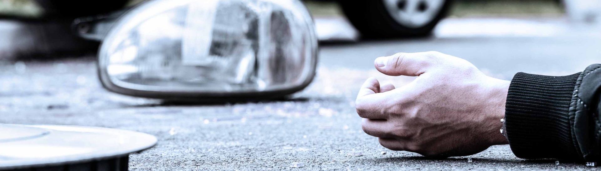 Close-up of a person's hand lying on the ground beside a smartphone, with a car headlight and part of a vehicle in the background, suggesting an accident or emergency