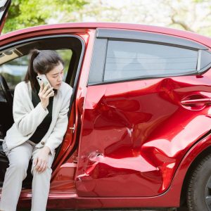 Woman sitting in a damaged red car while calling for help after a side-impact accident.