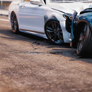 Two-car accident on a paved road, with a white car rear-ending a blue car, showing visible damage and fluid spill beneath both vehicles.