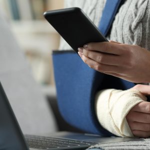 Close up portrait of a disabled woman with broken arm and sling using smartphone and laptop at home