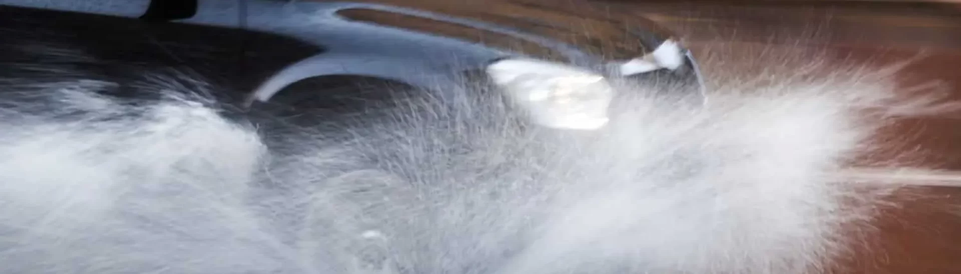 Car driving fast through a large puddle, splashing water dramatically to the side on a wet road.