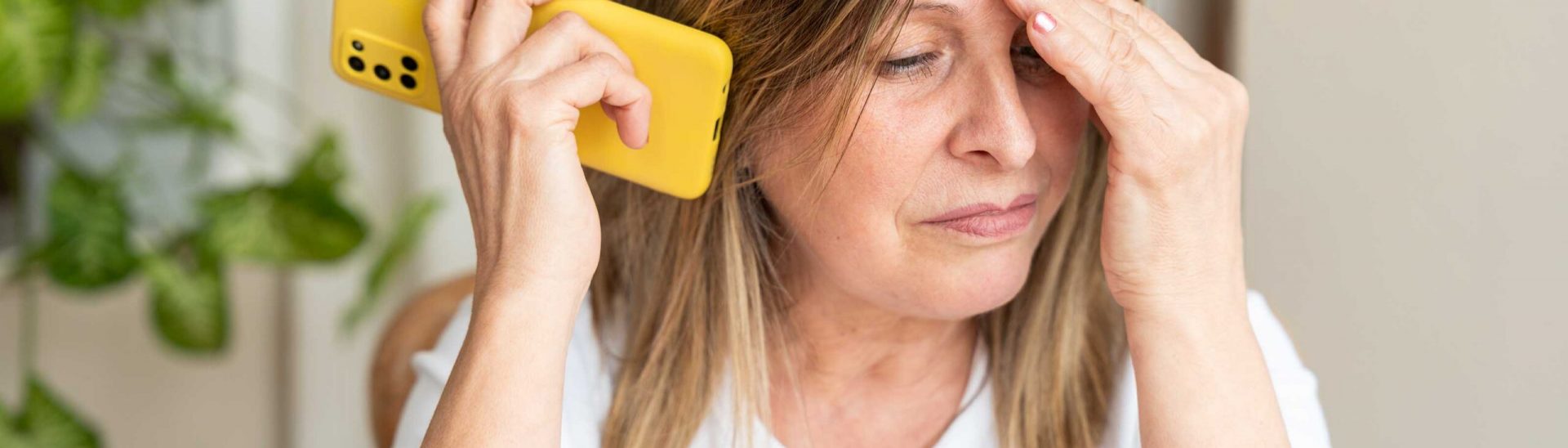 Mature female showing stress, touching forehead while listening intently to smartphone, displaying visible anxiety and emotional strain