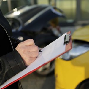 Person holding clipboard writing notes at the scene of a car accident, with damaged vehicles in the background, illustrating the importance of police reports in car accident injury claims in Arizona.