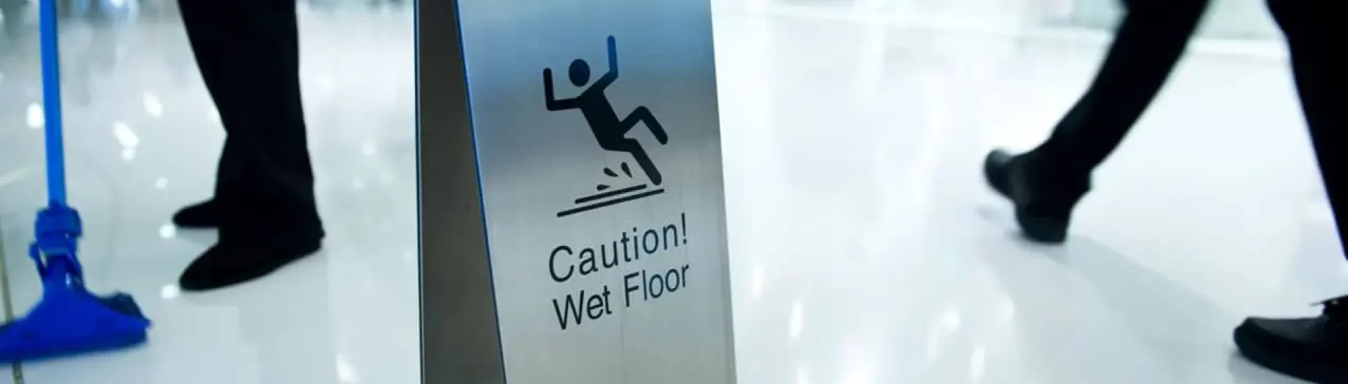 Warning sign on a shiny floor reading 'Caution! Wet Floor' with people walking nearby and a mop in the background.