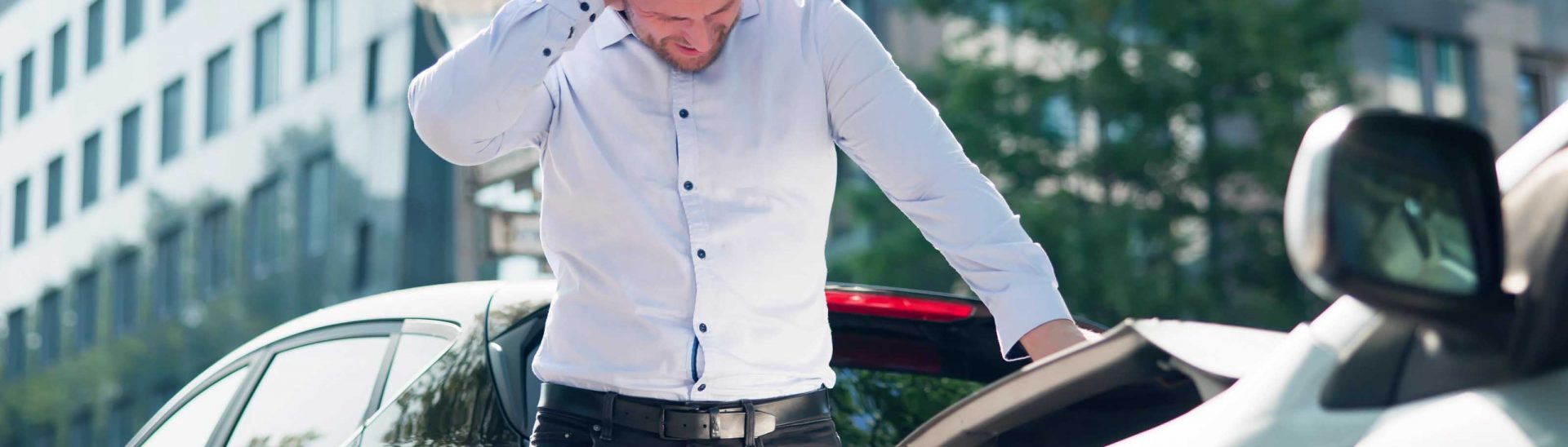 Man in a white shirt standing next to a damaged car, appearing stressed after a car accident, in an urban setting, emphasizing the need for immediate actions post-accident in Arizona.